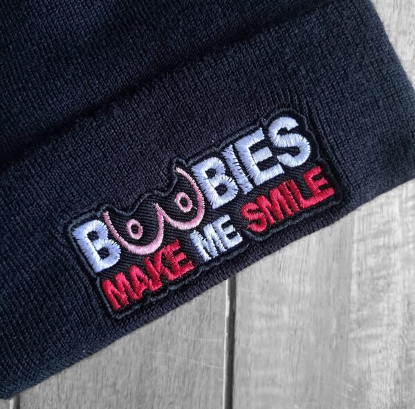 Close-up of the embroidered 'Boobies Make Me Smile' slogan on a black knit beanie