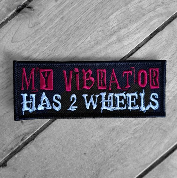 Rectangular sew-on patch reading ‘My Vibrator Has 2 Wheels’ in pink and white embroidered text on black background