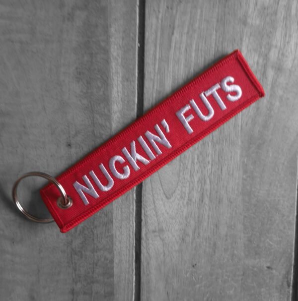 Red embroidered keytag with bold white “NUCKIN’ FUTS” text, shown on wooden surface (diagonal view)