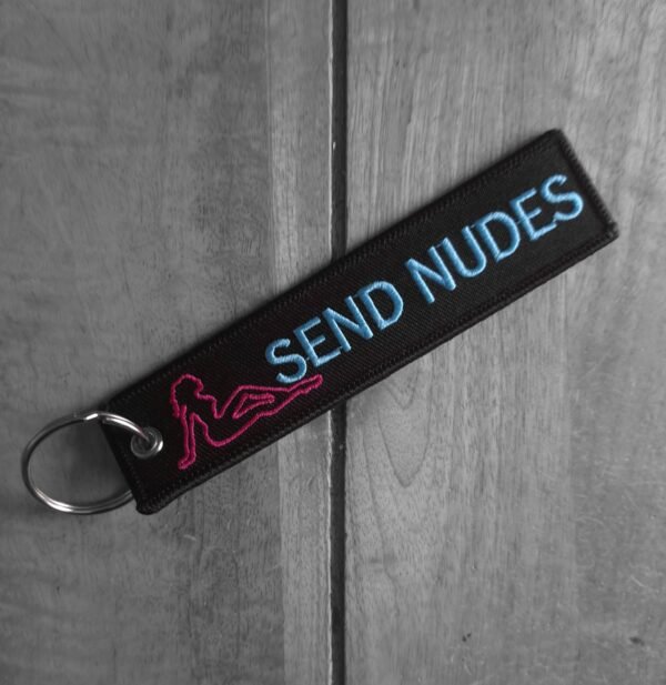 Embroidered keytag reading "SEND NUDES" in silver with pink pin-up silhouette, shown at a diagonal angle on wood.