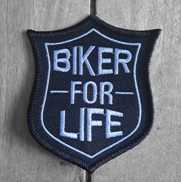 Straight-on view of “Biker for Life” embroidered patch in a shield shape with black background and light blue thread.