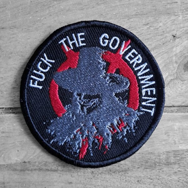 Front view of circular “Fuck the Government” patch with a shredded Guy Fawkes mask in grey and red on black background.