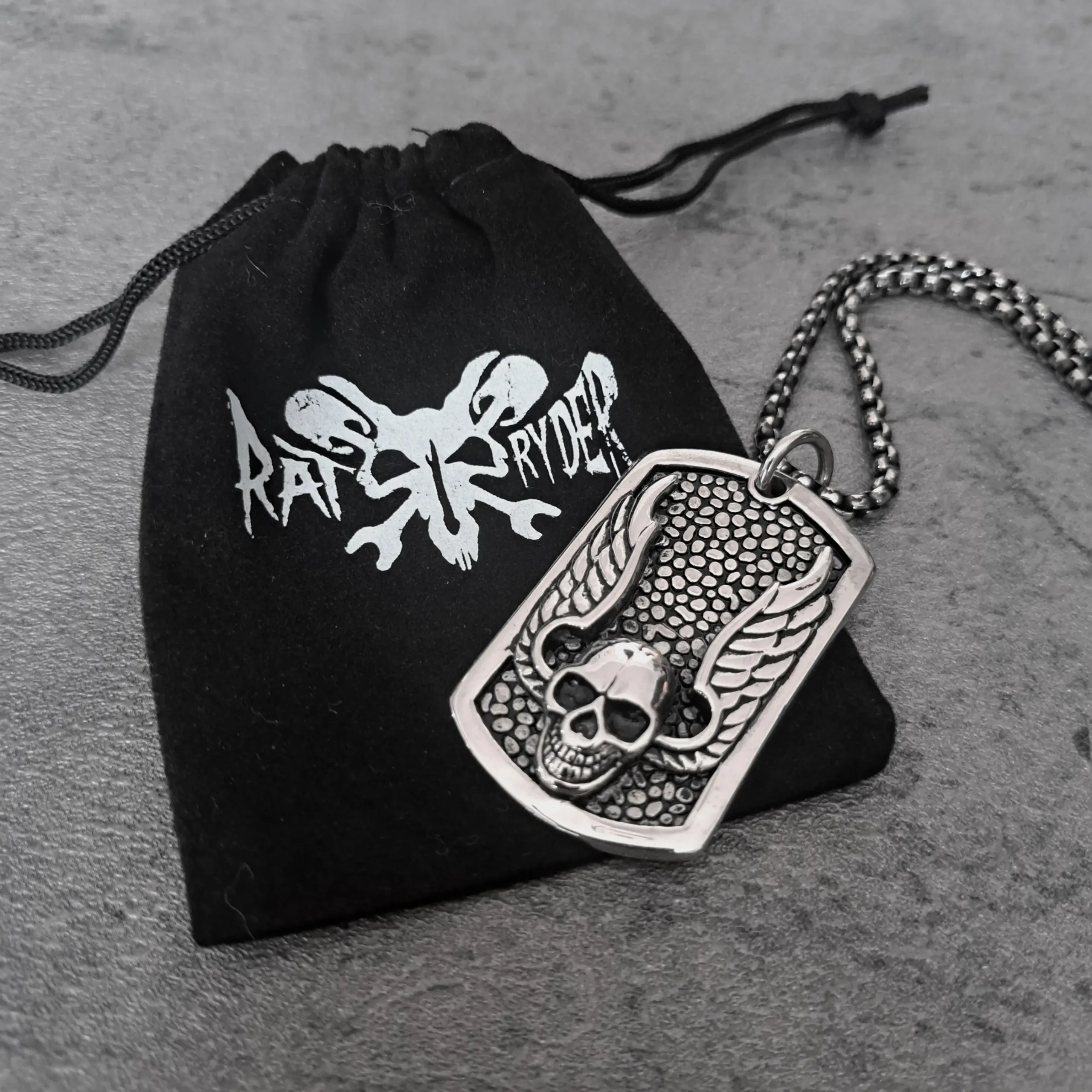 Winged Skull Dog Tag – Heavyweight 316L Stainless Steel Biker Pendant | RatRyder 3 Winged skull dog tag biker pendant resting on black RatRyder velvet gift bag